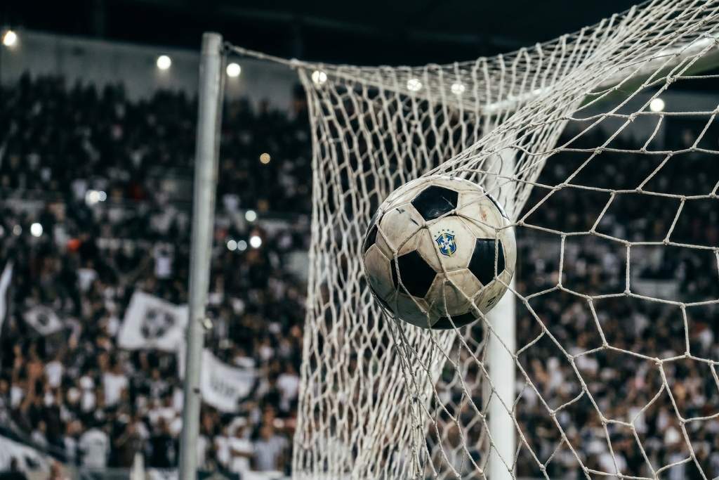Bola de futebol oficial com o escudo da CBF entrando no gol, presa nas redes brancas, durante uma partida noturna com estádio lotado ao fundo, ilustrando o momento de uma das maiores goleadas do futebol brasileiro.