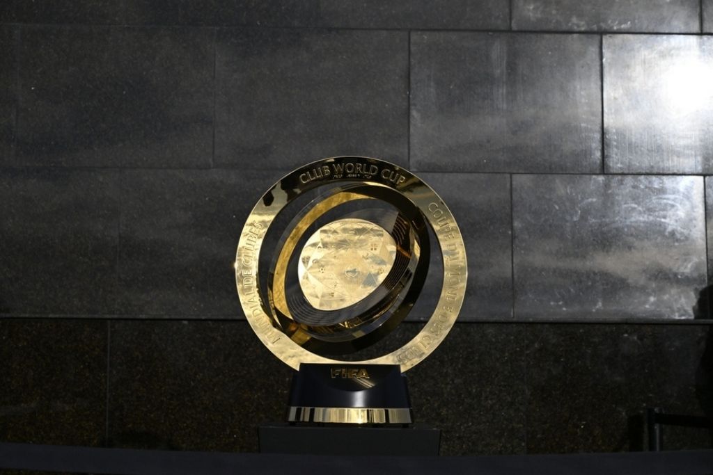 Troféu oficial da Copa do Mundo de Clubes da FIFA, competição disputada pelos clubes brasileiros campeões mundiais ao longo das décadas. O troféu dourado tem formato circular com anéis concêntricos e uma esfera central facetada, representando o globo terrestre. A base é preta com detalhes dourados e traz a inscrição "FIFA". Ao redor do anel externo estão gravadas as palavras "Club World Cup" e "Coupe du Monde des Clubs" em diferentes idiomas. O objeto está posicionado sobre uma superfície de granito escuro, diante de uma parede de pedra cinza.