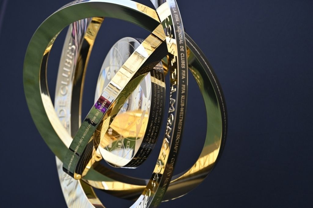 Fotografia em close do troféu do Mundial de Clubes da FIFA, o mesmo disputado pelos clubes brasileiros campeões mundiais ao longo das edições do torneio. A imagem destaca os detalhes da peça em ângulo aproximado, revelando os múltiplos anéis circulares dourados e prateados que se entrelaçam ao redor de uma esfera central facetada com reflexos metálicos. Nas faixas internas dos anéis estão gravadas inscrições em diversos idiomas com o nome da competição, sendo possível ler trechos como "Mundial de Clubes FIFA" e "FIFA Klub-Weltmeisterschaft". O fundo da imagem é azul escuro desfocado, o que realça o brilho e os reflexos dourados do troféu. A iluminação cria contrastes vibrantes entre as superfícies polidas da peça.
