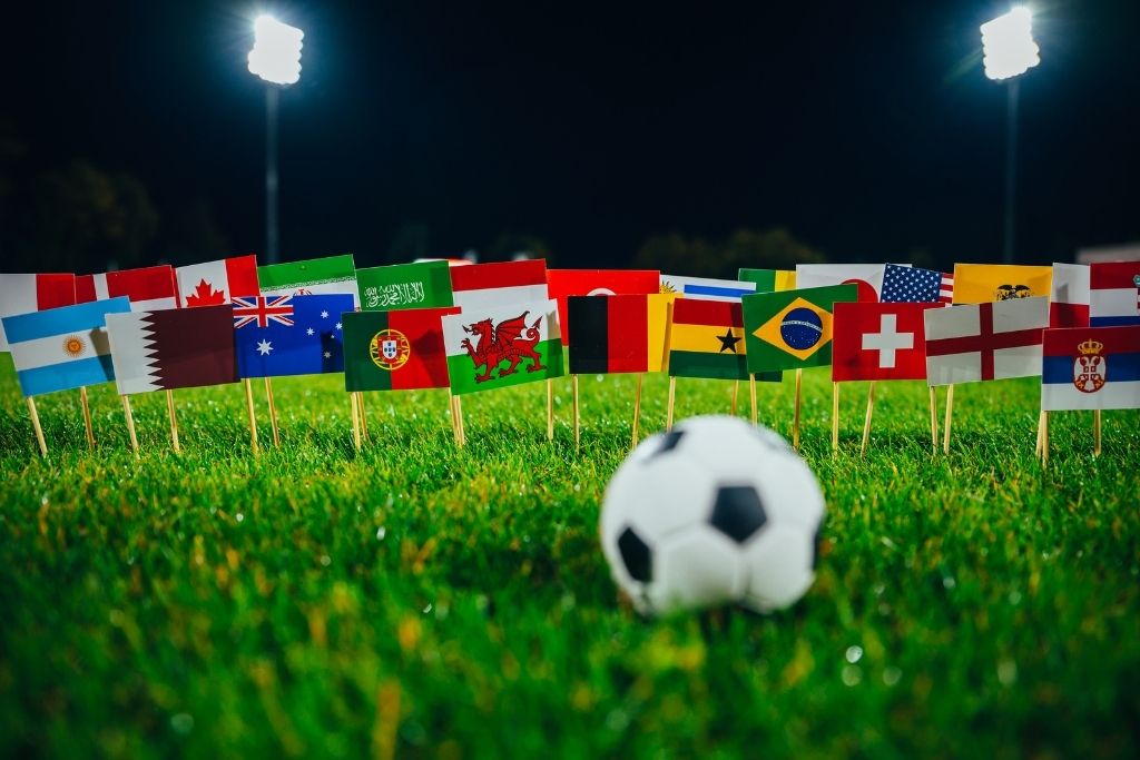 Bola de futebol no gramado iluminado à noite cercada por minibandeiras de diversas seleções participantes da Copa do Mundo, incluindo Brasil, Argentina, Portugal, Inglaterra e outras, simbolizando os países que figuram no ranking das seleções que mais perderam finais de Copa do Mundo.