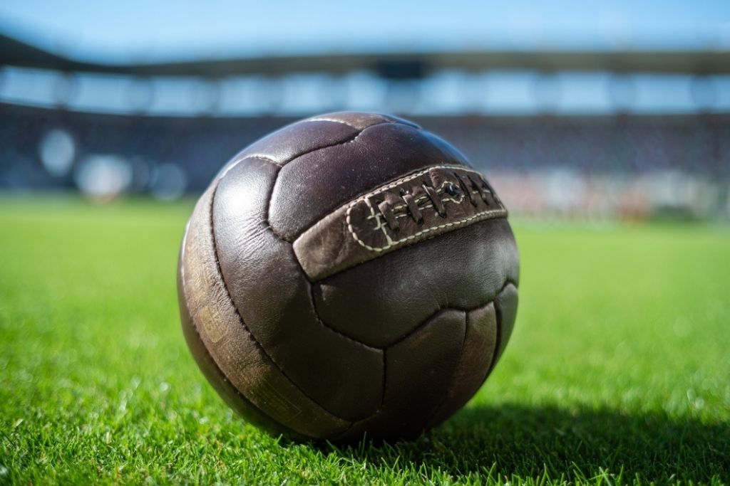 Uma bola de futebol de couro marrom com costuras grossas e aparentes, repousando sobre um gramado verde em um estádio desfocado ao fundo. A imagem ilustra o início e a história do futebol através de seus equipamentos clássicos.
