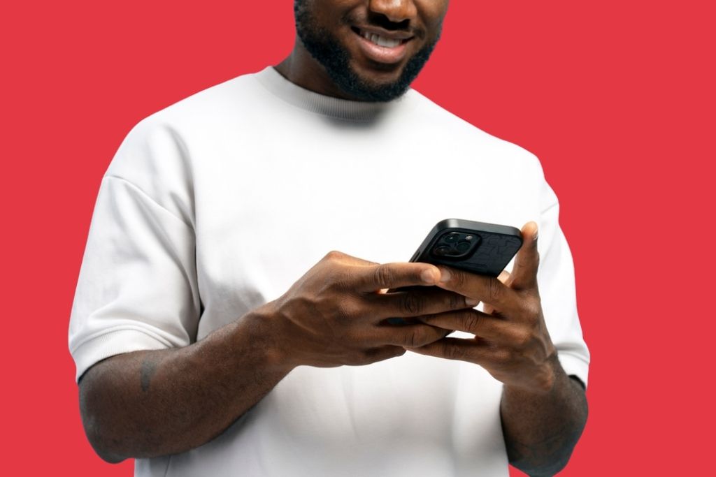 Homem sorridente de camiseta branca segurando um smartphone com as duas mãos, representando a experiência de uso do aplicativo BetMGM em um fundo vermelho sólido.