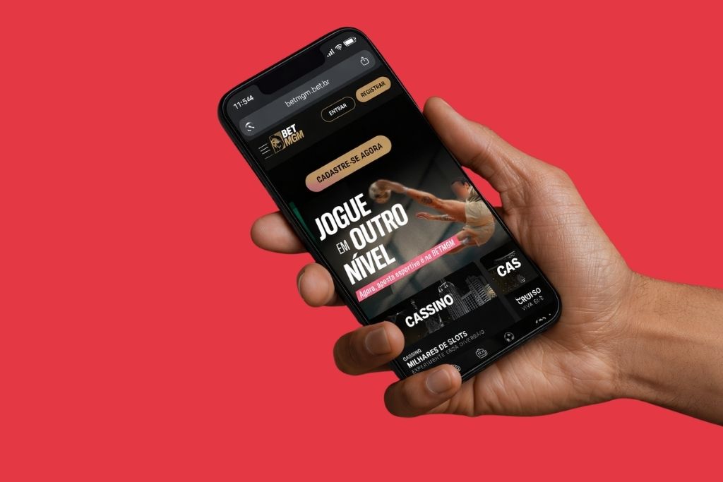 Uma mão segurando um smartphone que exibe a página inicial do site BetMGM. Na tela, destaca-se o botão dourado 'Registrar' no topo e um banner central com o texto 'Cadastre-se agora'.