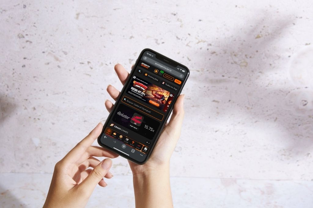 Uma pessoa segura um smartphone exibindo a interface móvel do site Aposta Ganha com destaque para o jogo Aviator e promoções de cashback em um fundo claro e minimalista.