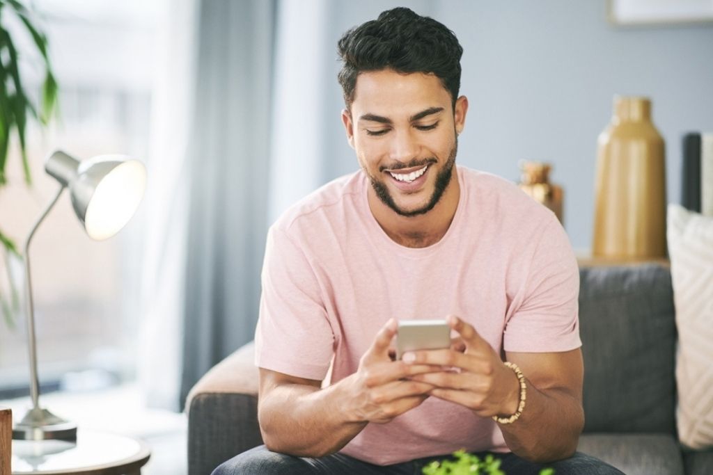 Um homem jovem e sorridente sentado em um sofá enquanto utiliza o celular para acessar as funcionalidades do aplicativo Aposta Ganha em um ambiente doméstico iluminado.