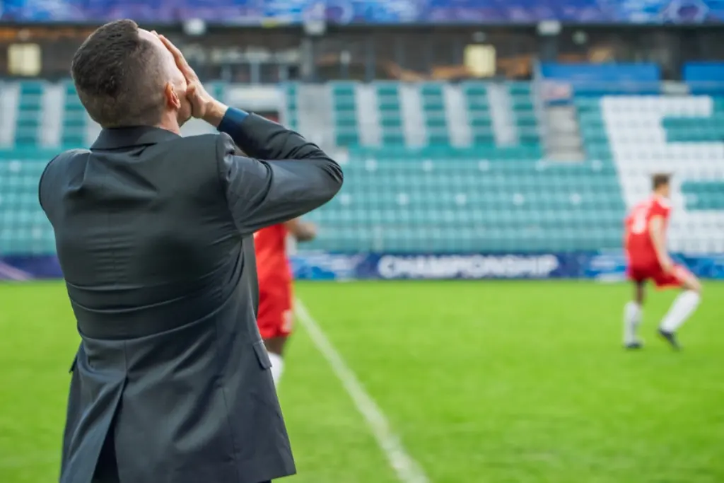Um treinador vestindo terno em um campo de futebol leva as mãos ao rosto em sinal de desespero enquanto observa o jogo ao fundo representando a frustração dos clubes rebaixados mais vezes no Brasileirão ao longo da história.