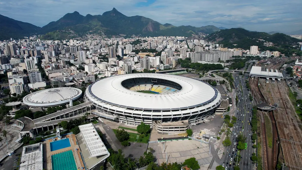Fotografia aérea diurna do complexo esportivo que recebeu os maiores públicos da história do Maracanã. A imagem mostra a icônica estrutura circular do estádio com sua cobertura branca, o ginásio Maracanãzinho ao lado, e o entorno urbano do Rio de Janeiro com montanhas e trilhos de trem ao fundo.