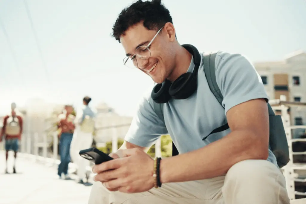 Homem jovem sorridente usando óculos e fones de ouvido, sentado ao ar livre e interagindo com seu celular. Ele parece satisfeito ao conferir resultados ou escolher as melhores bets para apostar.