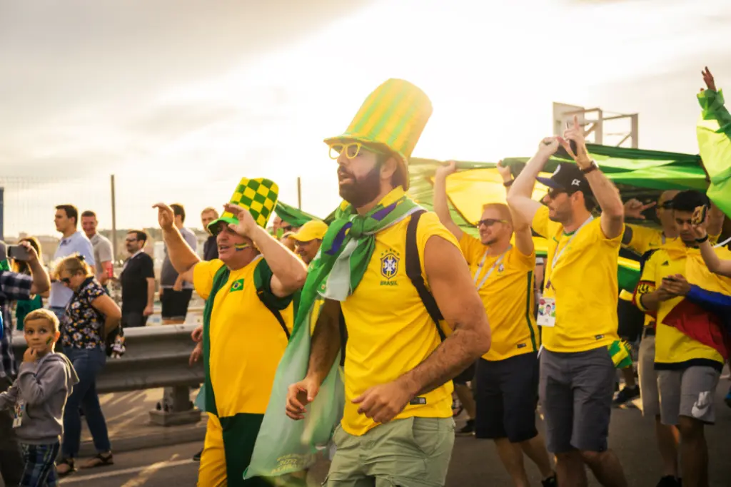 Grupo de torcedores brasileiros com camisas amarelas e acessórios verdes e amarelos caminha alegremente por uma rua durante o dia. Um homem em destaque usa um chapéu alto listrado e óculos coloridos, enquanto outros ao fundo carregam uma grande bandeira do Brasil. A imagem transmite a empolgação da torcida que se prepara para os horários dos jogos da Copa do Mundo 2026.
