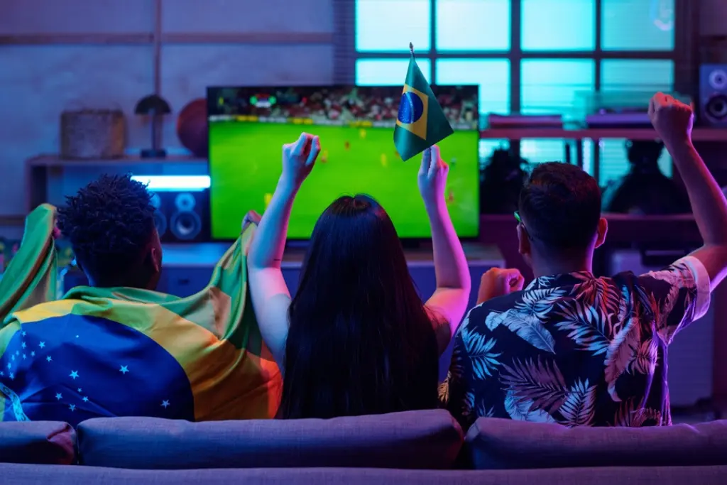 Três torcedores de costas — dois homens e uma mulher — sentados em um sofá assistindo à transmissão de um jogo da Seleção Brasileira em uma TV de tela grande. Eles celebram com os braços erguidos e seguram a bandeira do Brasil. O ambiente é uma sala com iluminação decorativa em tons de azul e rosa, ideal para acompanhar os horários dos jogos da Copa do Mundo 2026.