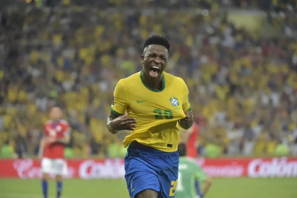 Vinícius Júnior, atacante da Seleção Brasileira, comemora um gol vibrando com a torcida durante uma partida importante para a tabela das eliminatórias da copa do mundo 2026. Ele veste a camisa amarela icônica em um estádio lotado.