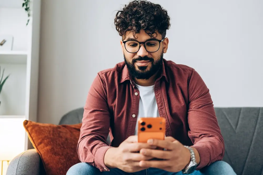Close de um homem jovem usando óculos e analisando dados no smartphone enquanto está sentado no sofá, focado em aprender como saber apostar no futebol com consciência.