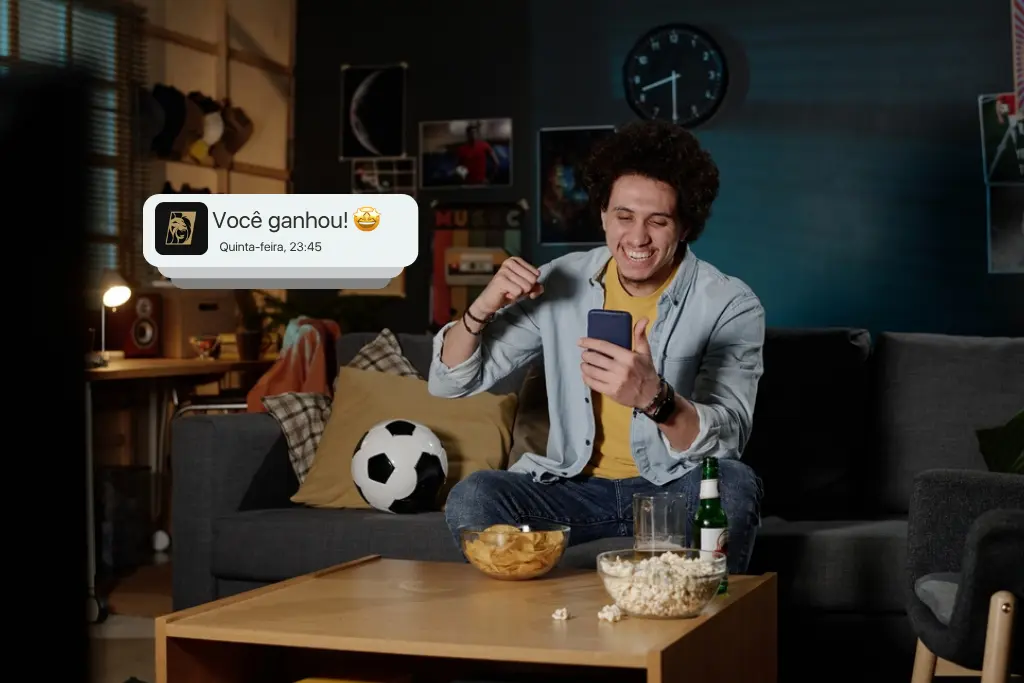 Homem jovem comemorando com o celular na mão ao receber notificação de vitória em dia de jogo, ilustrando como saber apostar no futebol de forma estratégica. No cenário, há uma bola de futebol e petiscos sobre a mesa.