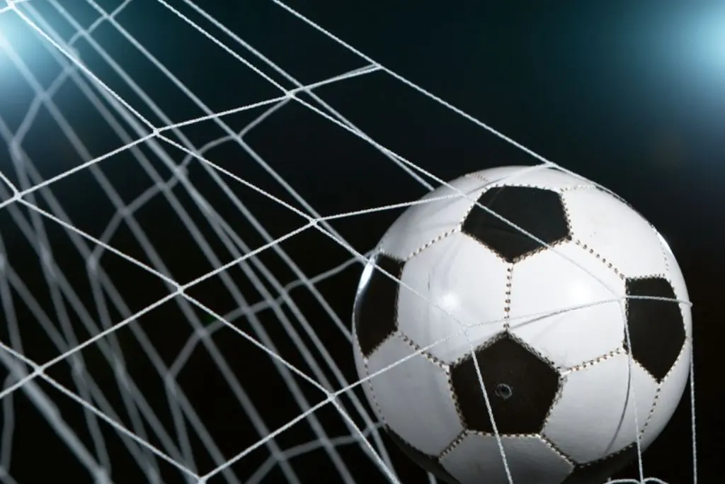 Close-up de uma bola de futebol clássica, branca e preta, estufando a rede após um gol sob as luzes de um refletor. A imagem ilustra o momento decisivo que contribuiu para a maior goleada da história do futebol brasileiro.