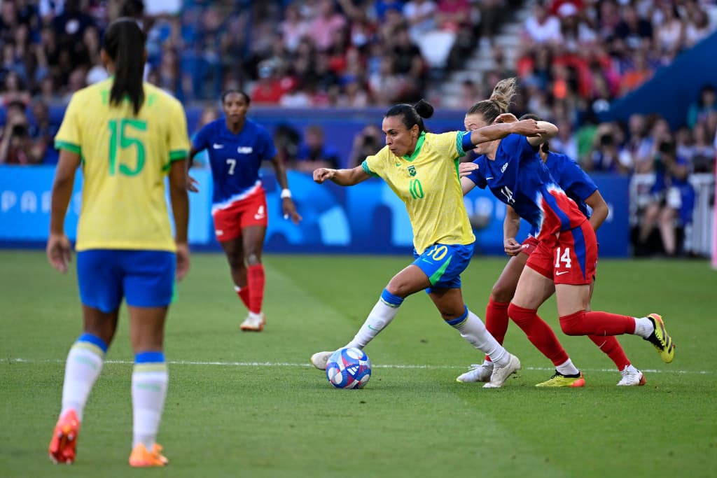 Registro fotográfico de um momento marcante na história do futebol feminino: a jogadora brasileira Marta, vestindo a camisa 10 da seleção, disputa a posse de bola contra duas defensoras dos Estados Unidos durante uma partida oficial em um estádio lotado. Marta aparece em movimento dinâmico de drible, simbolizando a técnica e a resistência da modalidade.