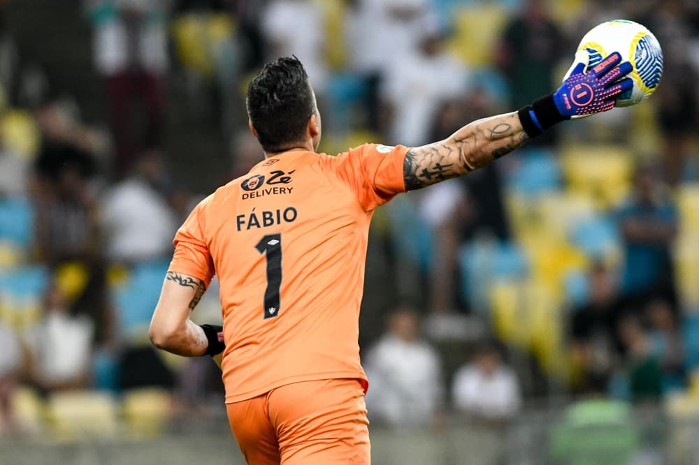 Registro do goleiro Fábio, reconhecido como o jogador com mais jogos na história do futebol, durante um momento de jogo. Ele veste camisa laranja e luvas profissionais.