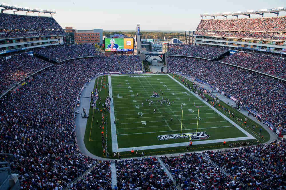 Vista aérea panorâmica de um grande estádio lotado durante uma partida da NFL, destacando o gramado verde demarcado com as linhas de jardas e o teto aberto. A atmosfera vibrante da torcida ilustra o ápice e o sucesso na história do futebol americano.
