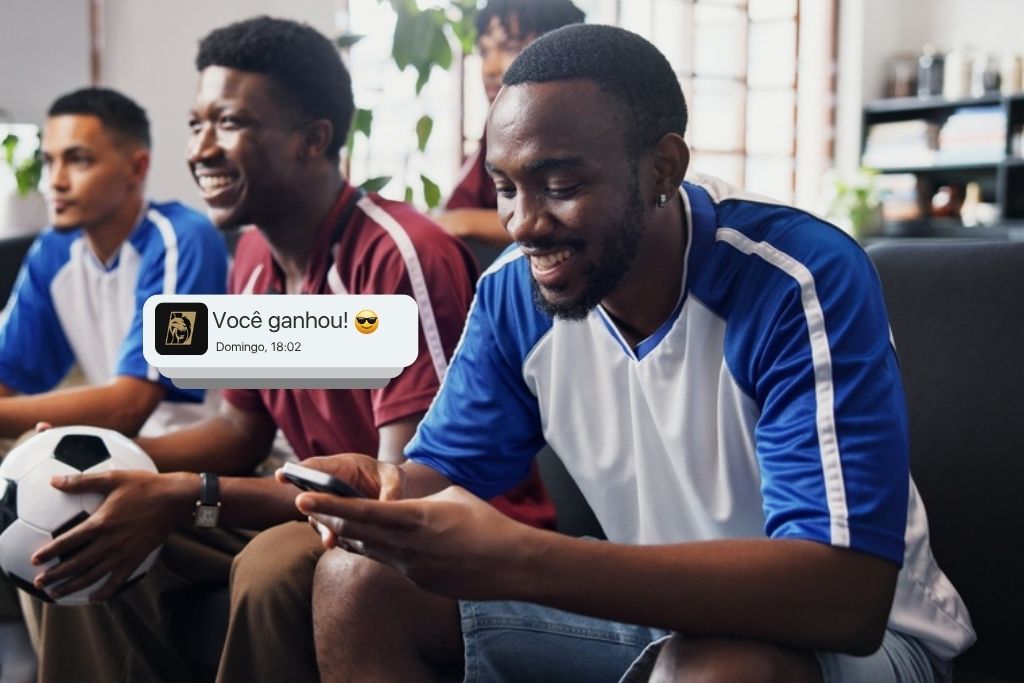Grupo de amigos assiste a um jogo reunido em um sofá. O homem em primeiro plano olha sorrindo para o celular, onde se destaca uma notificação flutuante com a mensagem "Você ganhou!". A cena demonstra a experiência de entretenimento e o resultado positivo de quem busca saber como apostar no futebol.