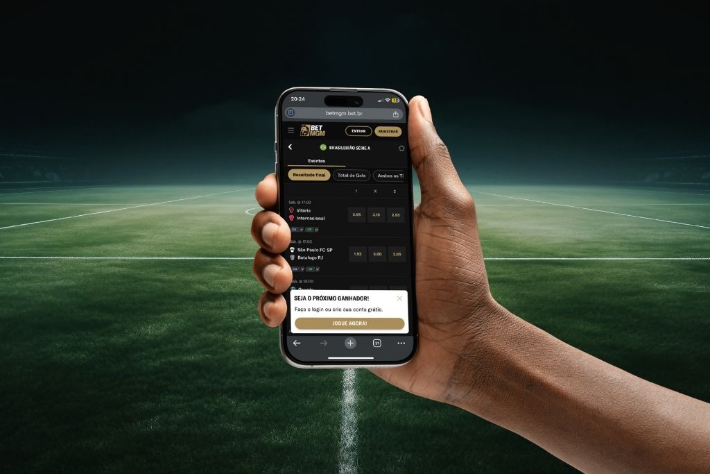 Uma pessoa segura um smartphone exibindo a interface de uma casa de apostas esportivas com jogos do Brasileirão Série A na tela. O fundo simula um campo de futebol desfocado, ideal para ilustrar tutoriais sobre como apostar no futebol pelo celular.