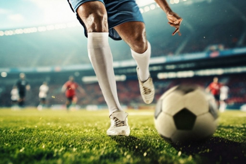 Imagem em plano detalhe e foco seletivo das pernas e pés de um jogador de futebol em movimento, vestindo calção azul, meias brancas e chuteiras claras, prestes a chutar uma bola clássica em um gramado verde. Ao fundo, o cenário desfocado mostra a atmosfera de um estádio lotado e iluminado, representando a paixão que consagra os top 10 melhores jogadores da história do futebol.