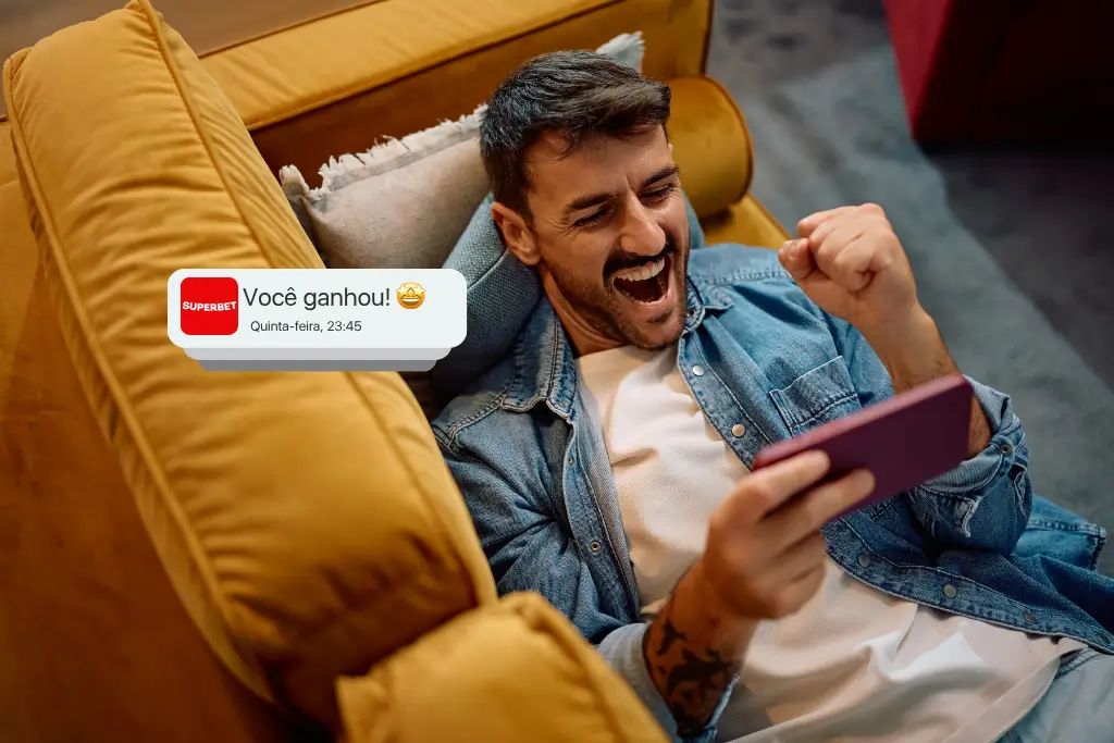 Homem comemorando enquanto olha para o celular deitado no sofá de casa, com notificação de vitória em aposta esportiva na tela; imagem ilustra o tema “como apostar no campeonato brasileiro”.