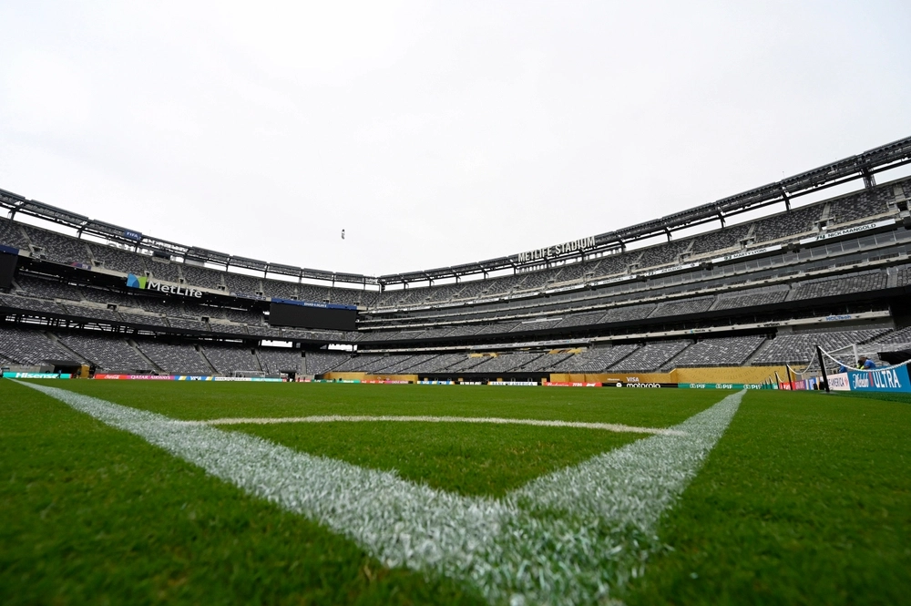 Fotografia de baixo para cima tirada do canto do campo de gramado artificial do MetLife Stadium, olhando em direção à arquibancada. A foto capta as linhas brancas do campo em primeiro plano, uma grande tela de vídeo e o nome "MetLife Stadium" no alto da arquibancada. O dia está nublado.