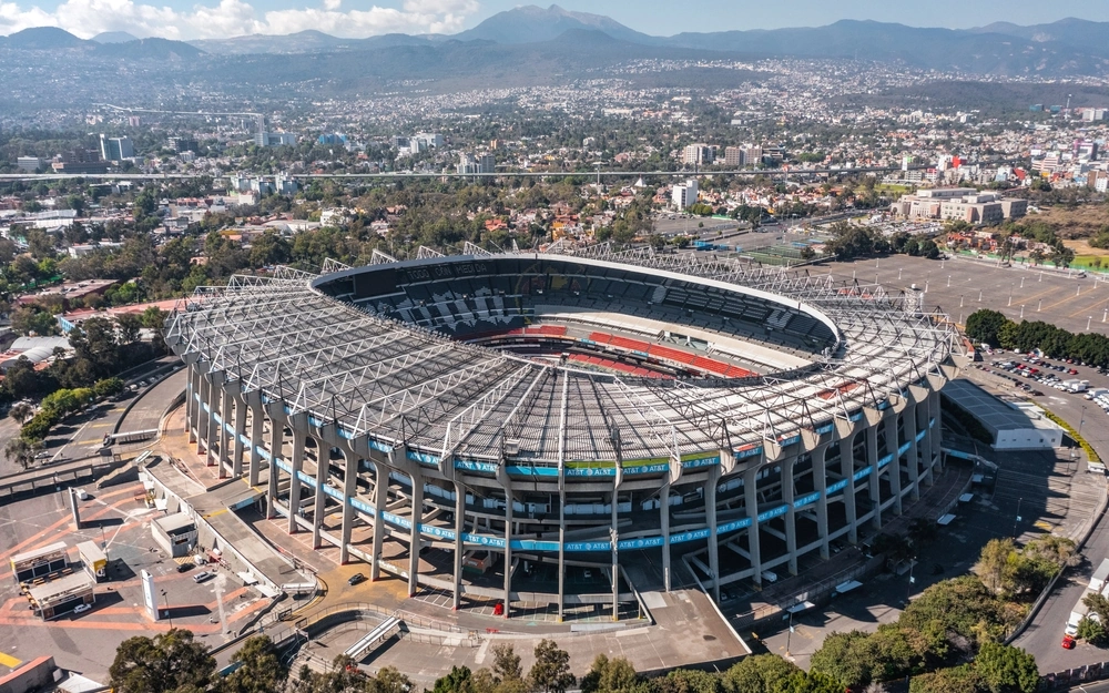 Fotografia aérea do Estádio Azteca, na Cidade do México. A imagem mostra o grande estádio ao ar livre com sua cobertura característica, cercado por uma área urbana densa e montanhas no horizonte sob um céu ensolarado.