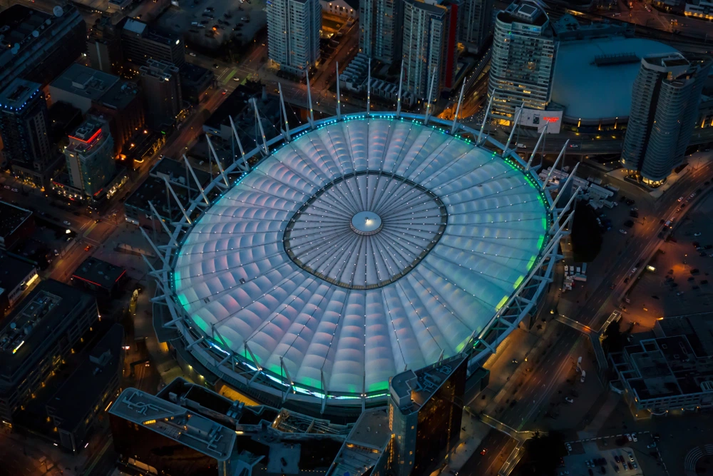 Fotografia aérea noturna do estádio BC Place, em Vancouver, Canadá. A imagem mostra o telhado de membrana retrátil do estádio iluminado com luzes azuis e verdes, com as luzes da cidade de Vancouver brilhando ao redor dele.