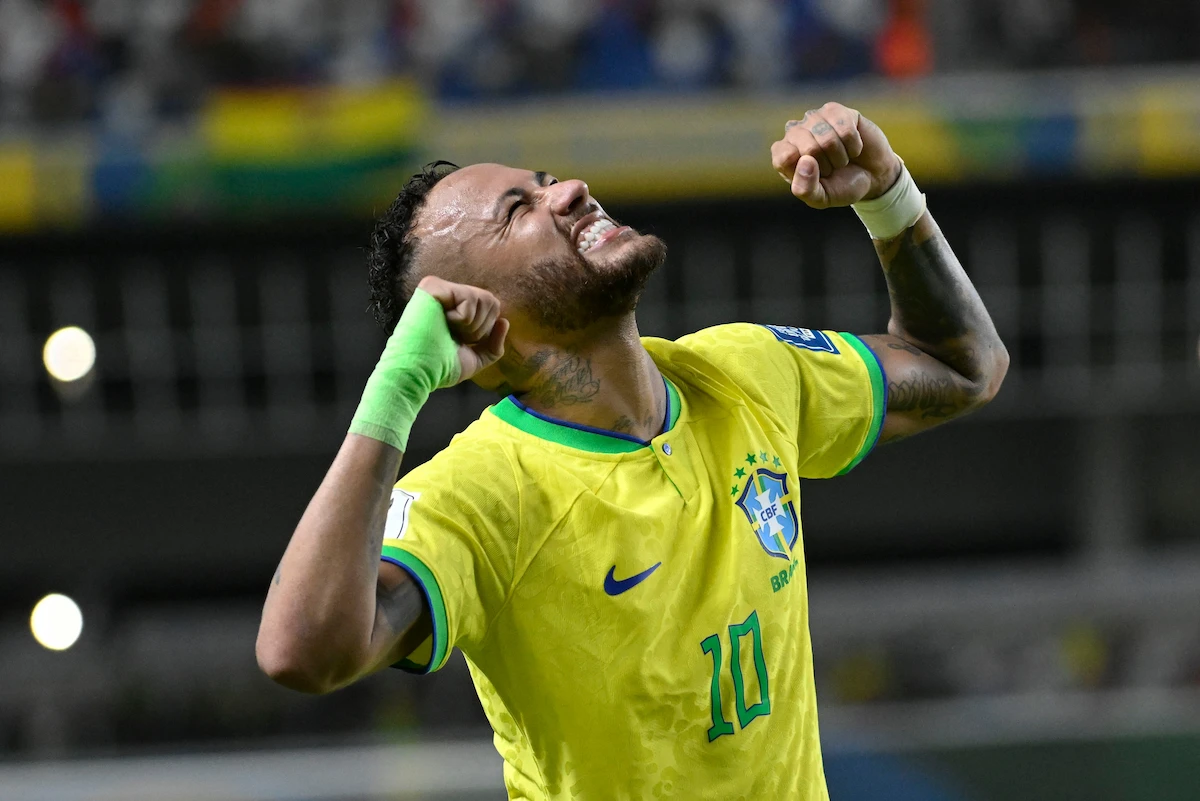 O jogador de futebol Neymar celebra intensamente em campo vestindo a tradicional camisa amarela número 10 da Seleção Brasileira. Ele aparece de perfil, olhando para cima com os olhos semicerrados e uma expressão de forte desabafo ou alívio, exibindo os dentes travados. Seus dois punhos estão cerrados e erguidos próximos ao rosto, com o pulso esquerdo envolto em uma faixa branca e o punho direito protegido por uma bandagem verde. O fundo da imagem mostra o ambiente desfocado de um estádio de futebol lotado durante uma partida. Imagens marcantes de jogadores como ele frequentemente ilustram os debates sobre as maiores polêmicas das convocações brasileiras em Copas do Mundo devido às grandes expectativas do público.