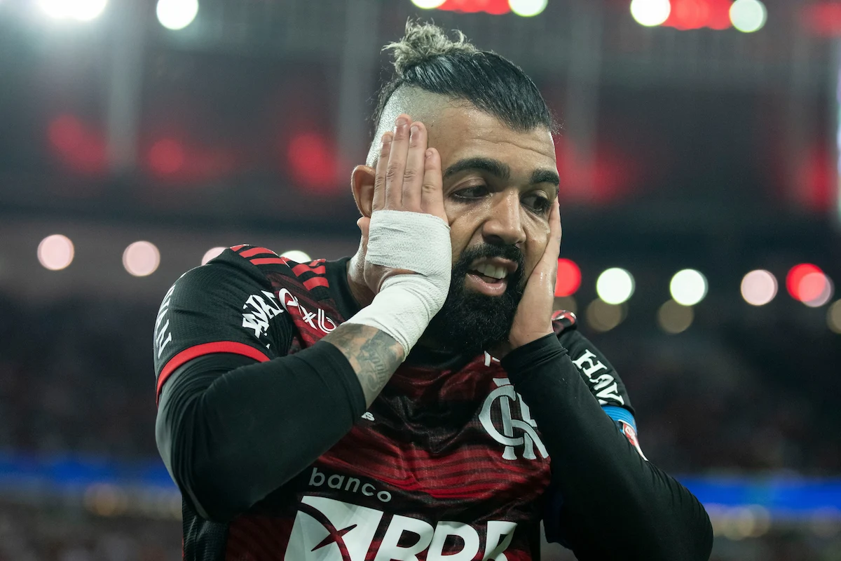 O atacante Gabigol é retratado em um momento de frustração ou incredulidade durante uma partida de futebol pelo Flamengo. Ele aparece vestindo a camisa rubro-negra do clube carioca, com as duas mãos espalmadas contra as bochechas e uma expressão de desânimo na face. Sua mão e pulso direitos estão protegidos por uma faixa médica branca. O jogador exibe seu característico penteado com cabelo amarrado no topo e as laterais raspadas, além de uma barba cheia. Ao fundo, as luzes desfocadas dos refletores do estádio criam um efeito de desfoque sob o céu noturno. A ausência de atletas de destaque do cenário nacional em torneios internacionais costuma figurar entre as maiores polêmicas das convocações brasileiras em Copas na visão dos torcedores.