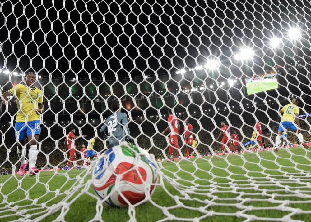 Previsões para copa do mundo 2026 ilustradas em uma cena de jogo com a bola dentro da rede após o gol, vista de dentro do gol com jogadores da Seleção Brasileira e adversários em campo sob refletores de um estádio lotado durante uma partida internacional.
