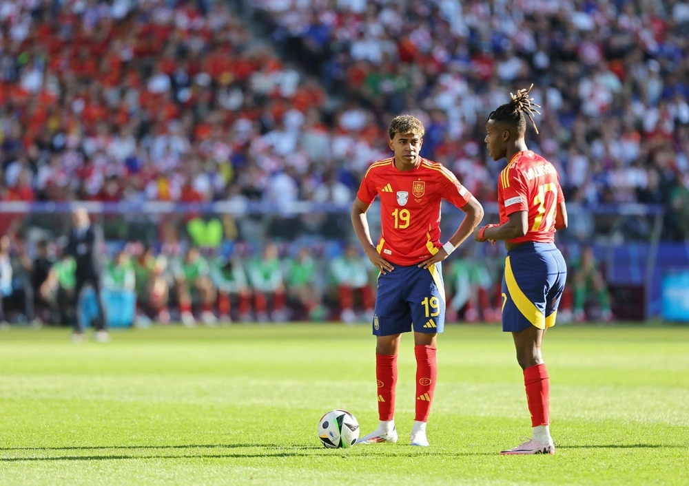 Dois jogadores da seleção espanhola (Lamine Yamal e Nico Williams) em campo durante uma partida de futebol, vestindo o uniforme vermelho e azul com detalhes em amarelo. Lamine Yamal, de frente com a camisa 19, apoia as mãos na cintura ao lado de Nico Williams, que está de costas com a camisa 17. Uma bola de futebol está no gramado à frente deles e a arquibancada ao fundo está lotada de torcedores. Imagem representando a Espanha na Copa 2026.
