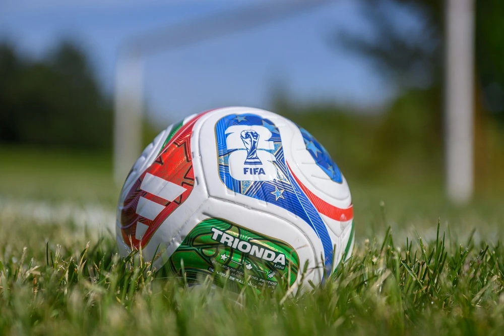 Uma bola de futebol oficial da FIFA no gramado, com as cores verde, vermelha e azul em destaque, ideal para quem planeja fazer uma aposta para Copa do Mundo. Ao fundo, as traves do gol aparecem desfocadas em um dia ensolarado.