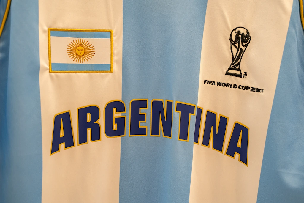 Detalhe em close da parte frontal de uma camisa listrada em azul-celeste e branco da Argentina na Copa 2026. Do lado esquerdo, destaca-se o patch oficial da FIFA World Cup 26; do lado direito, a bandeira do país com o Sol de Maio bordado em dourado. No centro, a palavra "ARGENTINA" está estampada em letras maiúsculas azuis com contorno amarelo.