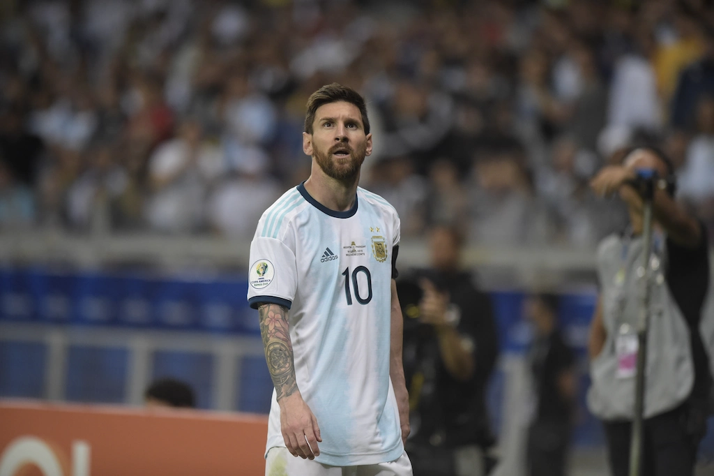 O jogador Lionel Messi em campo vestindo o uniforme oficial da seleção da Argentina na Copa 2026. Ele aparece de frente, com uma expressão focada olhando para cima, exibindo o número 10 no peito e suas tatuagens no braço direito, com o fundo do estádio e a torcida levemente desfocados.