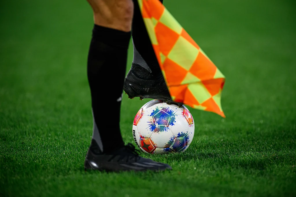 Close up em pés de árbitro assistente em um campo de futebol com um dos pés sobre uma bola, segurando uma bandeirinha. A pessoa usa meiões e chuteiras pretas.  