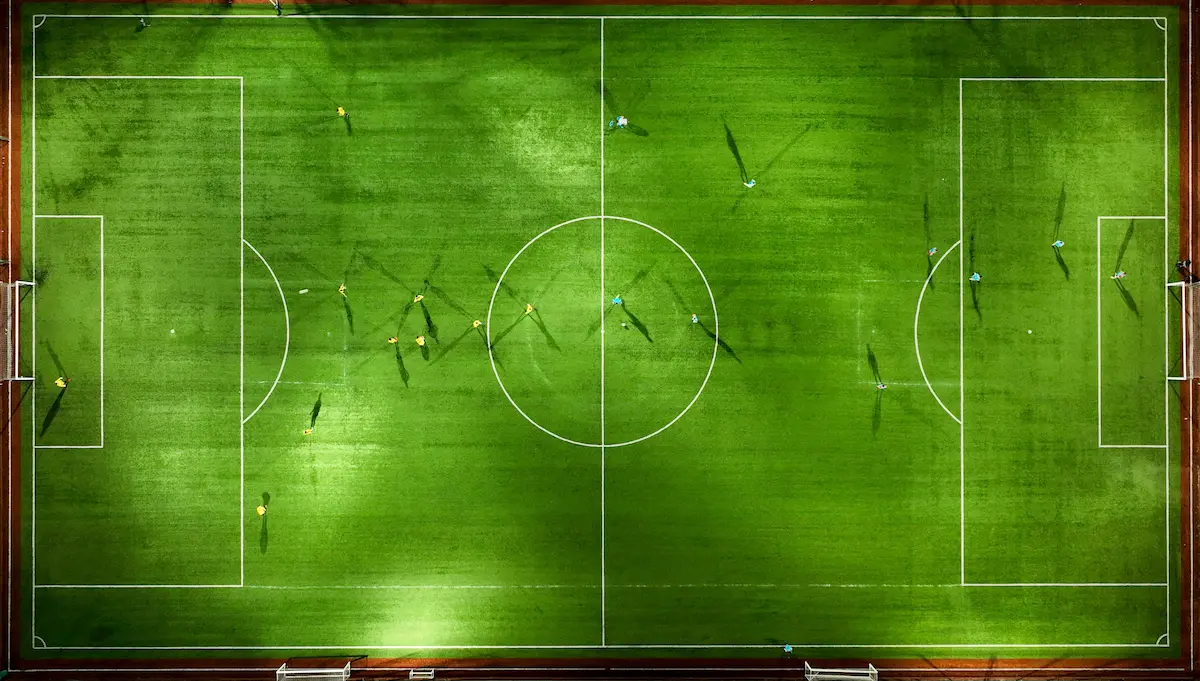Uma vista aérea de cima para baixo de um jogo de futebol jogado em um campo de futebol verde sob as luzes do estádio à noite. Os jogadores estão espalhados no campo