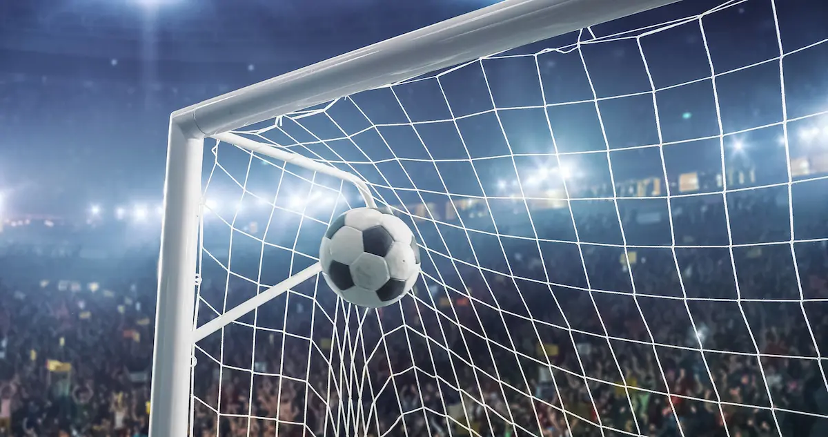 Fotografia em ângulo contra-plongé focando o canto superior interno de uma trave de futebol branca. Uma bola clássica preta e branca aparece estufando a rede no ângulo superior esquerdo, registrando o momento exato de um gol. Ao fundo, a arquibancada de um estádio lotado está desfocada sob os refletores acesos, criando uma atmosfera vibrante de jogo noturno.