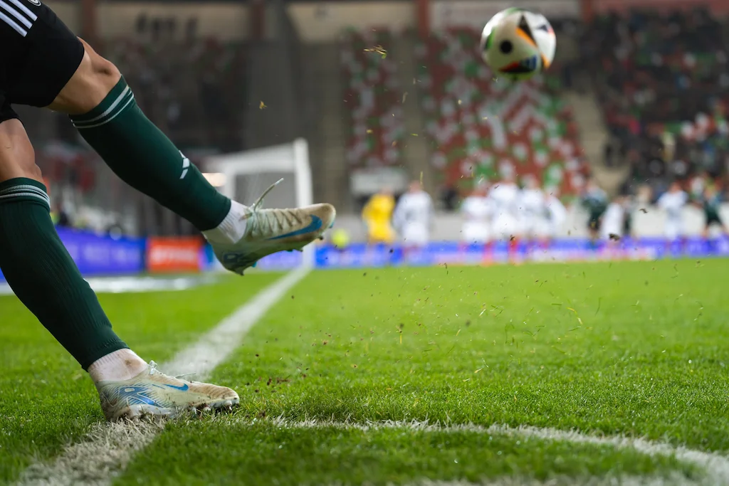 Close-up em ângulo baixo focado nas pernas e pés de um jogador de futebol no momento exato em que ele cobra um escanteio. O jogador veste meiões verdes e chuteiras brancas com detalhes em azul