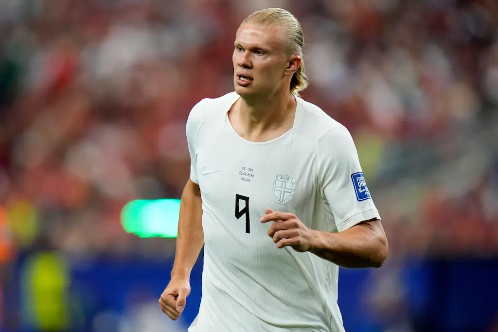 Erling Haaland, jogador da seleção da Noruega, em campo usando uniforme branco, com arquibancada desfocada ao fundo.